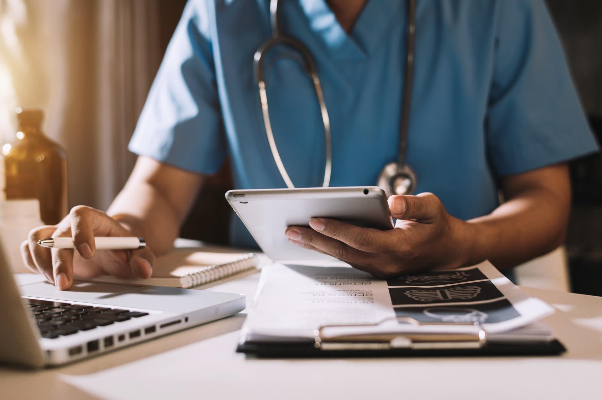 A medical professional sits at a desk reviewing information on a tablet while typing on a silver laptop holding a pen.